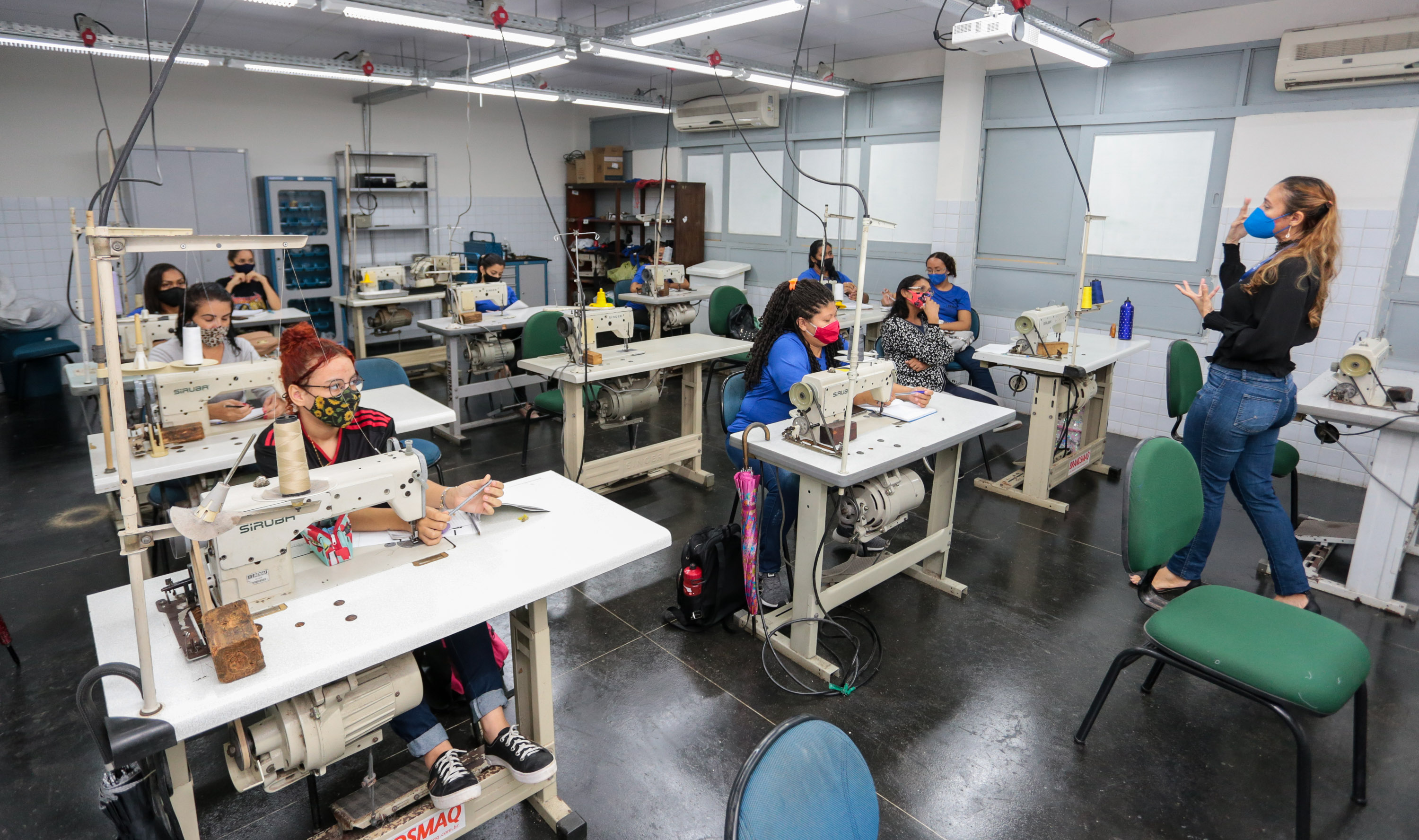 sala de aula de um curso de costura, as alunas estão sentadas em mesas com máquinas de costuras